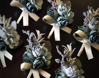 May include: Several floral boutonnieres with blue and green fabric flowers, cream-colored ribbons, and sprigs of dried foliage. The boutonnieres are arranged on a dark surface, showcasing their delicate details and color palette.