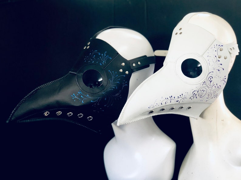 Puede incluir: Dos m&aacute;scaras de m&eacute;dico de la peste, una negra con detalles azules y otra blanca con detalles morados. Ambas m&aacute;scaras tienen un pico largo y agujeros para los ojos. Las m&aacute;scaras est&aacute;n expuestas en cabezas de maniqu&iacute;.