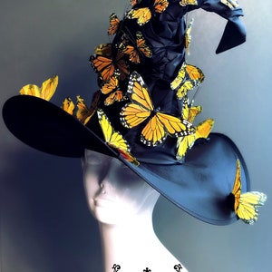 May include: A black witch hat with a tall, pointed crown adorned with numerous yellow and black butterfly embellishments. The hat is worn on a white mannequin head. The text "MASQUERADE STORE .COM" is visible at the bottom of the image.