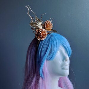 May include: A gold headband with pine cone and twig accents. The headband is worn by a mannequin with pink and blue hair.