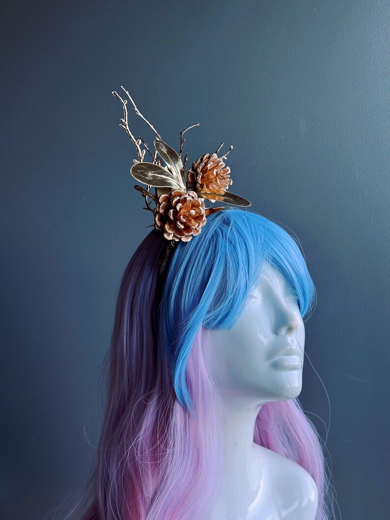 May include: A gold headband with pine cone and twig accents. The headband is worn by a mannequin with pink and blue hair.