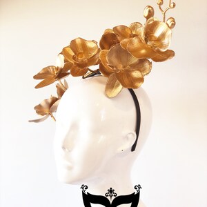 May include: Gold orchid flower headband. The headband is black and the flowers are gold. The headband is worn on a white mannequin head.