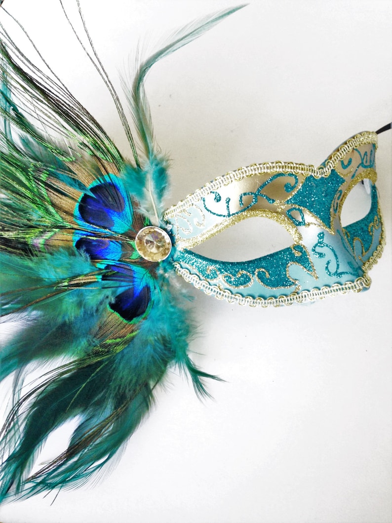 Masquerade mask for women in gold and turquoise color with matching peacock, turquoise and teal shade feathers attached to the right hand side of the mask. A round gem sits on the side of the mask where the feathers are attached as a finishing touch.
