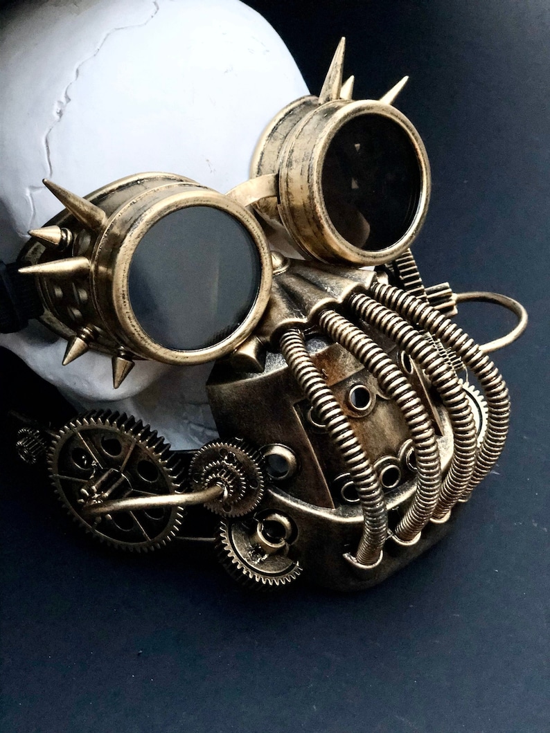 May include: A gold steampunk mask with goggles and gears. The mask has a skull-like design with a pair of round goggles and a series of gears and pipes attached to the side.