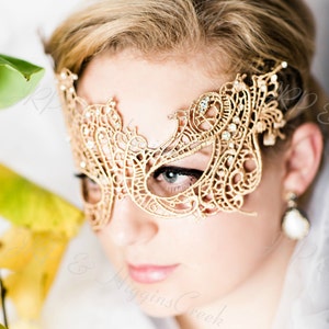 May include: A woman wearing a gold lace masquerade mask with rhinestones. The mask covers her eyes and part of her forehead. She is wearing a white dress and has blonde hair.