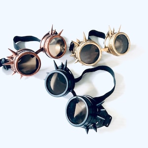 May include: Four pairs of steampunk goggles with spiked detailing. The goggles have round lenses and adjustable black straps. The colours are bronze, gold, and black. The design has a vintage, industrial aesthetic.