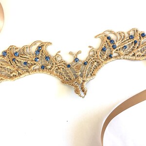 May include: Gold-coloured lace crown with blue gemstones. The crown features an intricate, openwork design and is adorned with small, round blue gemstones. A tan ribbon is attached.