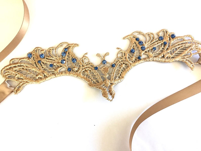 May include: Gold-coloured lace crown with blue gemstones. The crown features an intricate, openwork design and is adorned with small, round blue gemstones. A tan ribbon is attached.