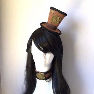 Op de afbeelding: Een bruine hoge hoed met zwarte trim en gouden tandwielen, gedragen als een hoofdband. De hoed zit op een mannequin met lang zwart haar en een bruine leren choker met een gouden tandwielbedel.
