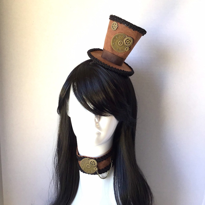 Op de afbeelding: Een bruine hoge hoed met zwarte trim en gouden tandwielen, gedragen als een hoofdband. De hoed zit op een mannequin met lang zwart haar en een bruine leren choker met een gouden tandwielbedel.