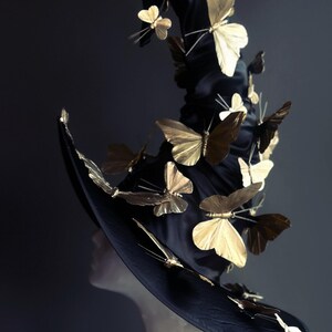 May include: A black witch hat adorned with numerous gold-colored butterfly embellishments. The hat has a wide brim and a tall, pointed crown. The text "MASQUERADE STORE" is visible on the upper right corner.