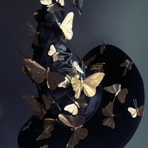 May include: A black witch hat adorned with numerous gold-colored butterfly embellishments. The butterflies are of varying sizes and appear to be attached to the hat, creating a whimsical and elegant design. The hat has a wide brim and a tall, pointed crown.