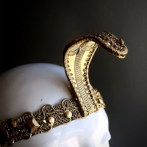 May include: A gold-colored cobra crown, featuring intricate filigree and geometric embellishments. The snake's head is raised, showcasing detailed scales and a curved design. The crown is set on a white mannequin head against a dark background.