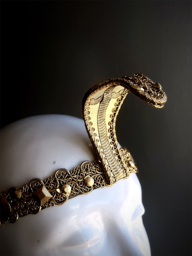 May include: A gold-colored cobra crown, featuring intricate filigree and geometric embellishments. The snake's head is raised, showcasing detailed scales and a curved design. The crown is set on a white mannequin head against a dark background.