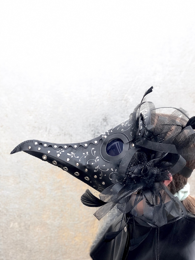 May include: A black leather plague doctor mask with silver accents and a black feather trim. The mask has a long beak and a round eye piece.