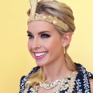 May include: A woman wearing a gold-colored cobra headpiece and a matching headband. The woman is also wearing a dark blue dress with gold and white embellishments. The background is a solid yellow.