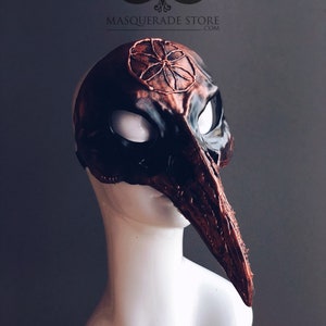 Pu&ograve; includere: Una maschera da ballo personalizzata in nero e rame con un lungo becco curvo. La maschera presenta un design dettagliato sulla fronte, che ricorda un fiore o una stella stilizzata. La maschera &egrave; esposta su una testa di manichino bianca. "Custom Witche's Mark!" &egrave; scritto in nero nella parte inferiore dell'immagine.