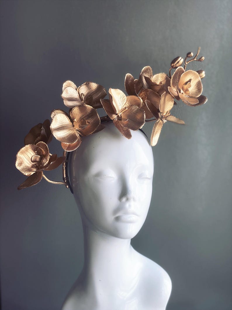 May include: A gold headband with a cluster of gold orchid flowers. The headband is worn on a white mannequin head.
