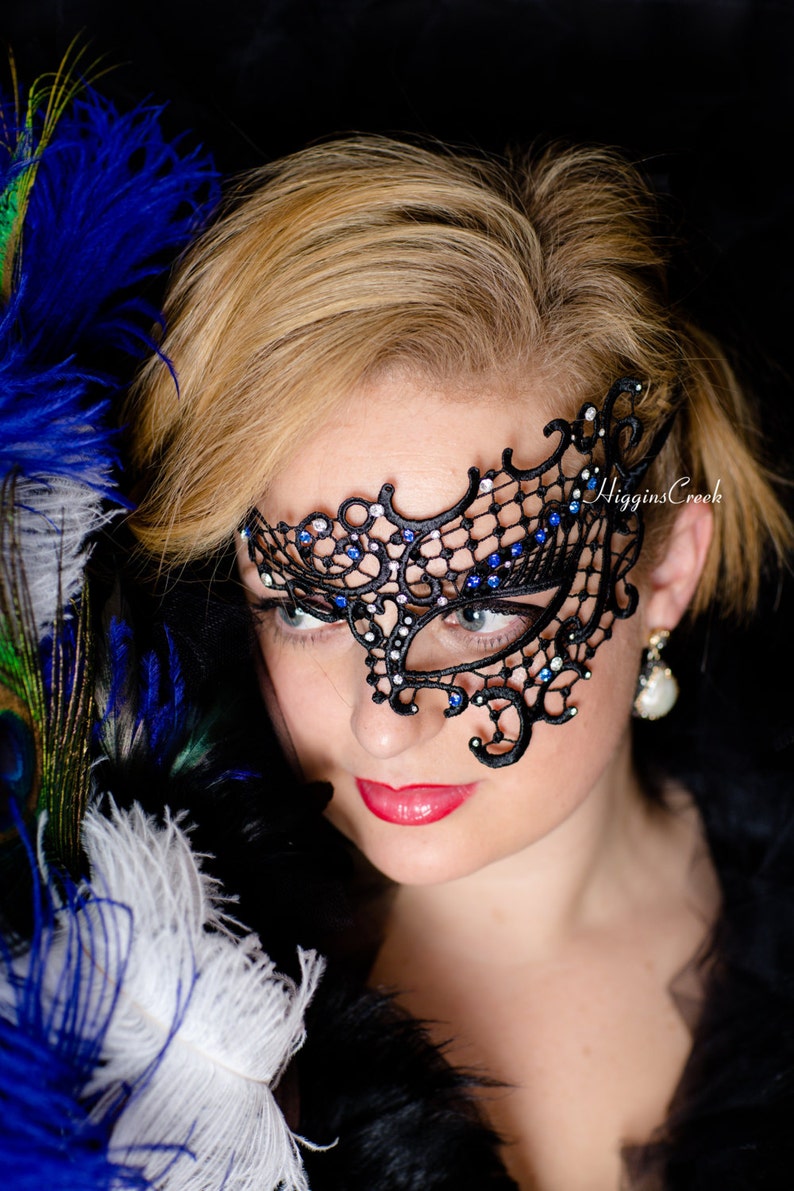 Peut inclure: Une femme portant un masque de bal de dentelle noire avec des strass bleus. Le masque couvre son &oelig;il droit et une partie de son front. Elle porte une tenue noire et a les cheveux blonds.