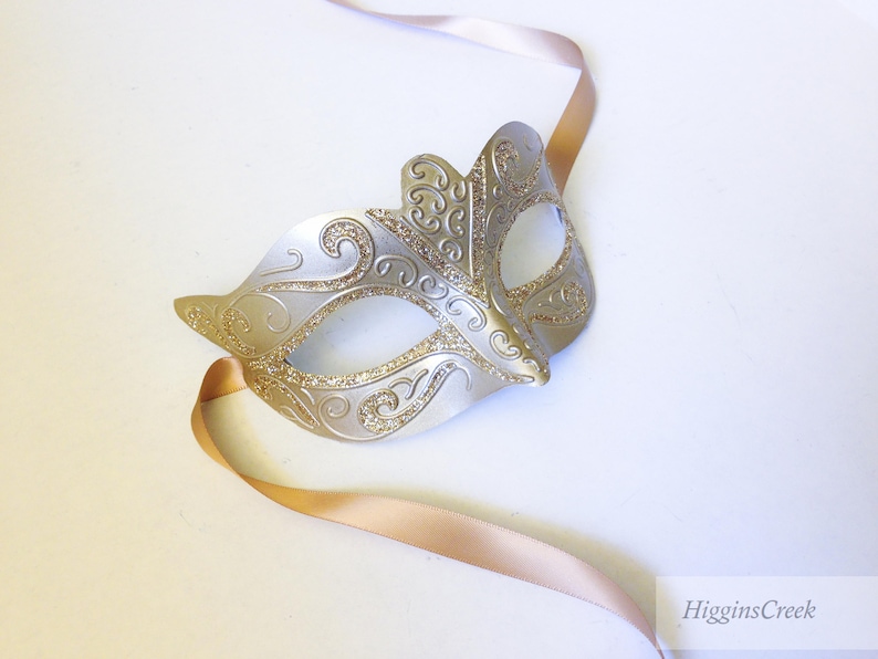May include: A silver and gold masquerade mask with intricate details and a gold ribbon attached.