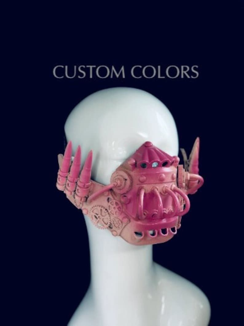 May include: A pink and tan steampunk mask with detailed gear designs and a textured surface. The mask has tubes, spikes, and a central dome. The text "CUSTOM COLORS" is displayed above the mask.