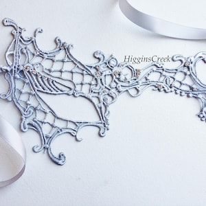 May include: A silver lace masquerade mask with rhinestones. The mask is tied with a silver ribbon.