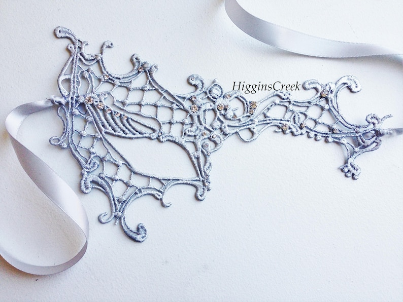 May include: A silver lace masquerade mask with rhinestones. The mask is tied with a silver ribbon.