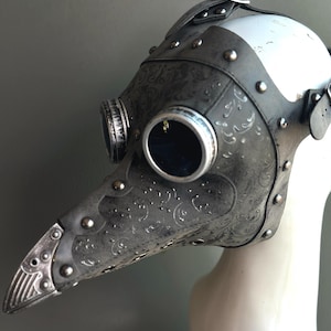 May include: A steampunk-style plague doctor mask in shades of gray and silver. The mask features a long, beaked nose, round eye lenses, and decorative embossed patterns. Rivets and straps add to the vintage aesthetic. The mask is displayed on a white mannequin head.