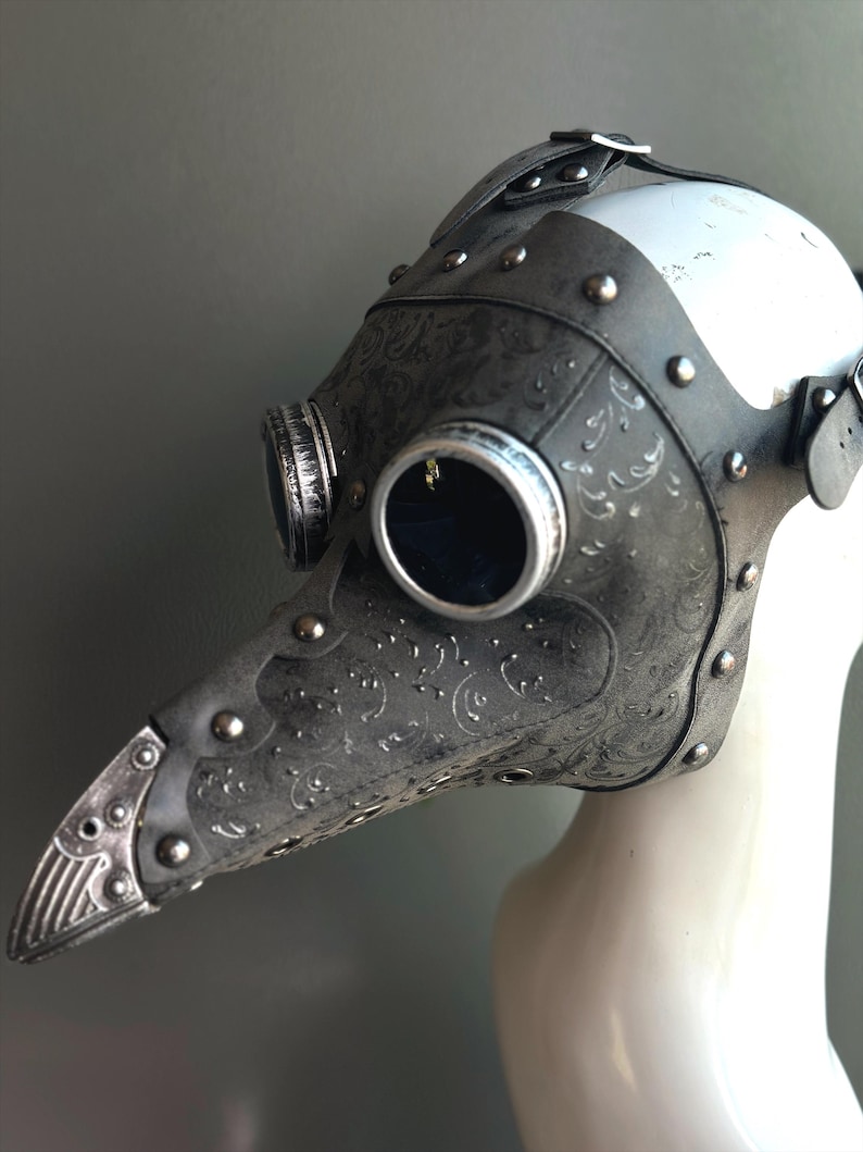 May include: A steampunk-style plague doctor mask in shades of gray and silver. The mask features a long, beaked nose, round eye lenses, and decorative embossed patterns. Rivets and straps add to the vintage aesthetic. The mask is displayed on a white mannequin head.