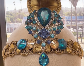 Peut inclure: Une couronne de mascarade dorée ornée de bijoux turquoise et ambre. La couronne présente un grand pendentif en forme de goutte d'eau turquoise et un groupe central de pierres précieuses turquoise et bleues plus petites. La base de la couronne est ornée de filigranes dorés.