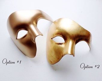 Könnte beinhalten: Zwei Masken, eine goldfarben und eine in einem helleren Goldton. Beide Masken haben ein einzelnes Augenloch und eine geschwungene Form.