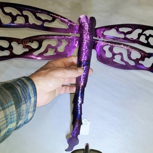 May include: Purple metal dragonfly wall art with intricate cutouts. The dragonfly has a long, slender body and two pairs of wings.