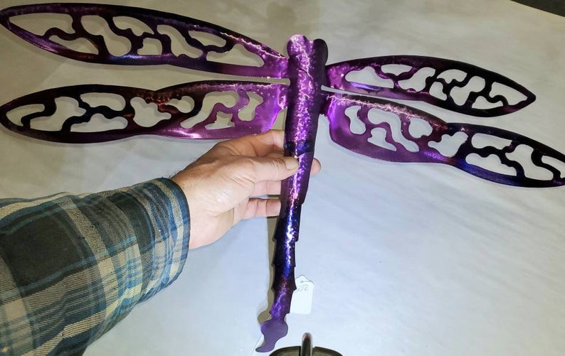 May include: Purple metal dragonfly wall art with intricate cutouts. The dragonfly has a long, slender body and two pairs of wings.