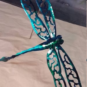 May include: A teal blue metal dragonfly with intricate cut-out designs on its wings. The dragonfly is mounted on a thin metal rod.