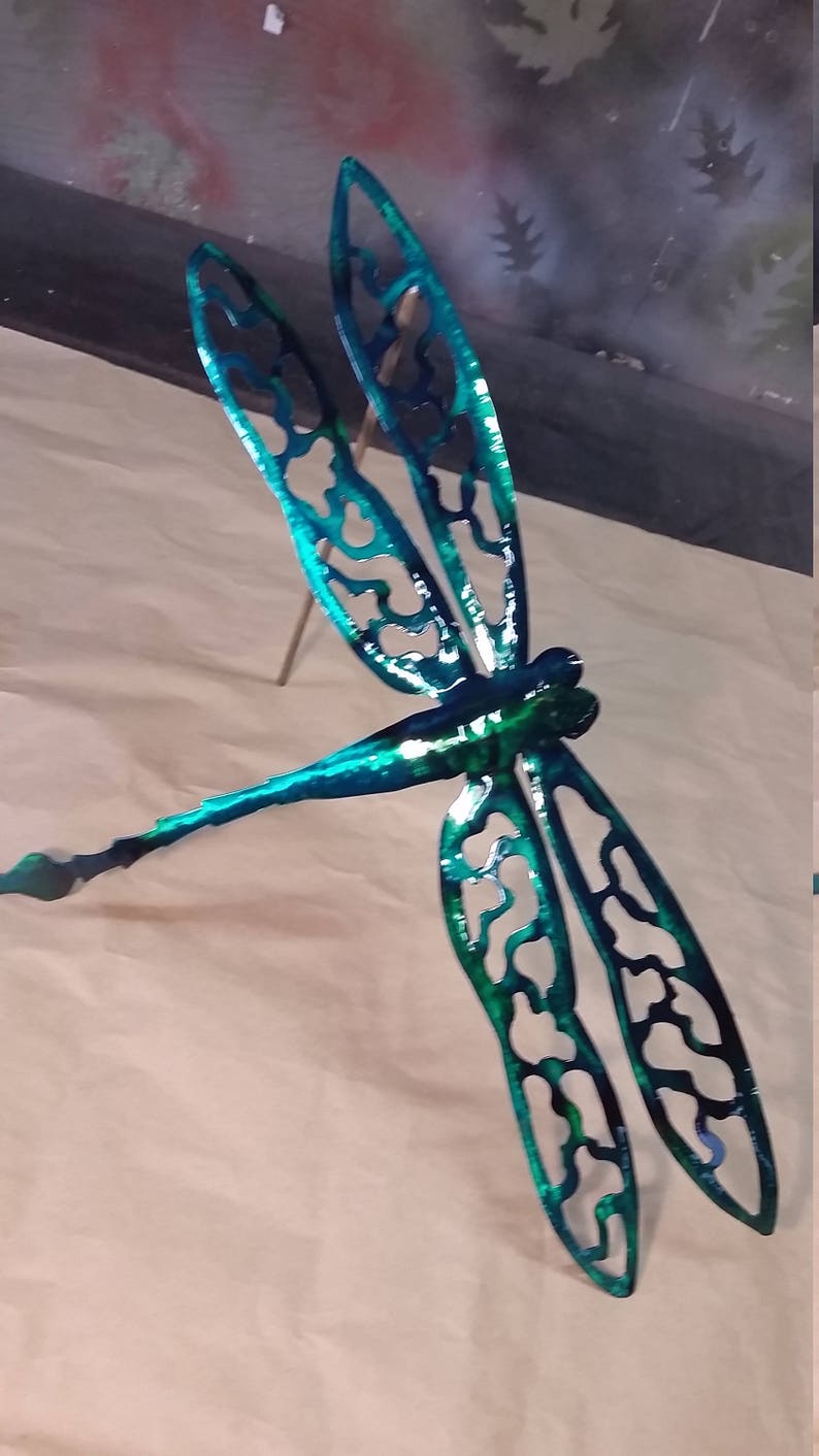 May include: A teal blue metal dragonfly with intricate cut-out designs on its wings. The dragonfly is mounted on a thin metal rod.