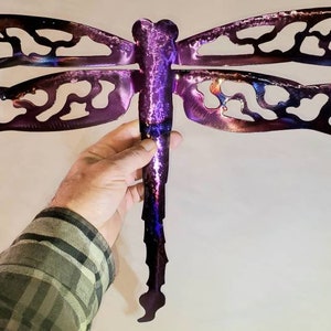 May include: A purple metal dragonfly wall art with a cut-out design. The dragonfly has a long, thin body and two pairs of wings.