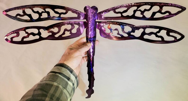 May include: A purple metal dragonfly wall art with a cut-out design. The dragonfly has a long, thin body and two pairs of wings.