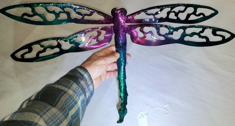 May include: A metal dragonfly wall art with a green, purple, and blue ombre finish. The dragonfly has intricate cutouts in its wings.
