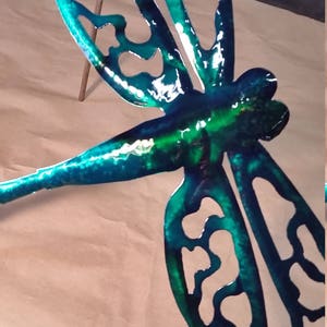 May include: A teal blue metal dragonfly with intricate cutouts and a green iridescent finish. The dragonfly is mounted on a wooden stick.