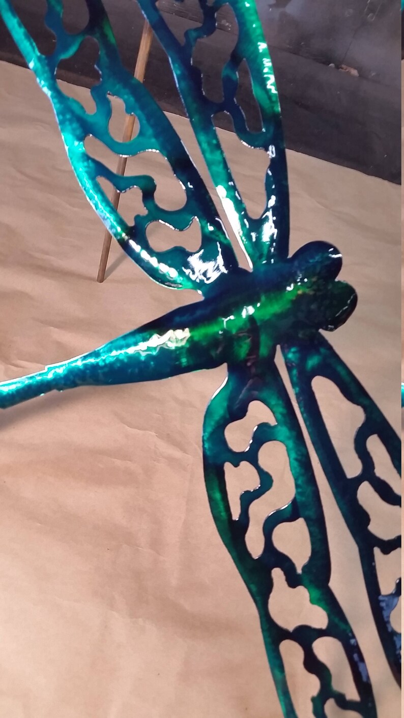 May include: A teal blue metal dragonfly with intricate cutouts and a green iridescent finish. The dragonfly is mounted on a wooden stick.