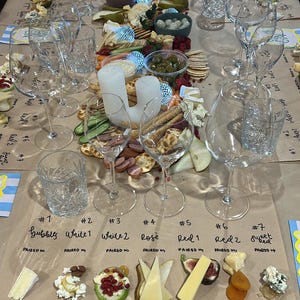 Puede incluir: Una mesa preparada para una cata de vinos y quesos, con copas de vino, queso, galletas saladas y otros acompañamientos. La disposición incluye varios tipos de queso, frutas y frutos secos, con etiquetas manuscritas que indican los maridajes.