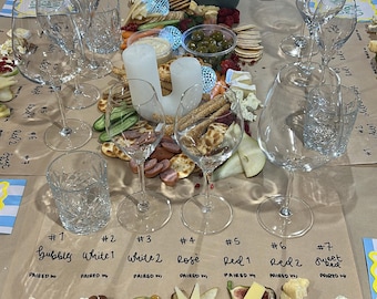 May include: A table setting featuring wine glasses, cheese, crackers, and various accompaniments. The arrangement includes multiple types of cheese, fruits, and nuts, with handwritten labels indicating pairings. The setup suggests a wine and cheese tasting event.