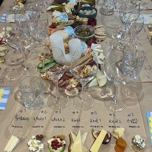 Puede incluir: Una mesa preparada para una degustación, con copas de vino, queso, galletas saladas y otros acompañamientos. La disposición incluye diferentes tipos de queso, frutas y frutos secos, con etiquetas manuscritas que indican los maridajes. La configuración sugiere un evento de cata de vinos y quesos.