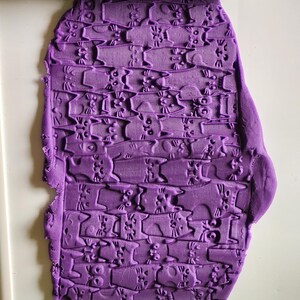 May include: A lime green cylindrical rolling pin with a repeating cat pattern embossed on its surface, positioned above and below a flat, purple sheet of modeling clay that displays the same cat pattern pressed into its texture.