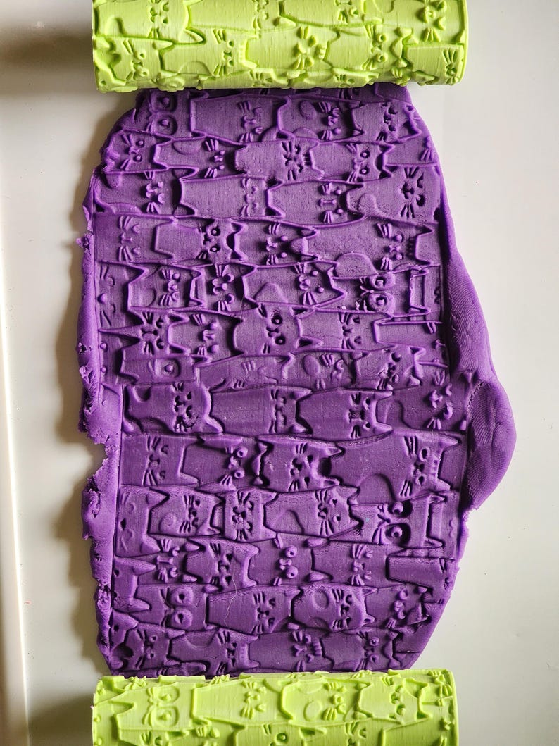 May include: A lime green cylindrical rolling pin with a repeating cat pattern embossed on its surface, positioned above and below a flat, purple sheet of modeling clay that displays the same cat pattern pressed into its texture.