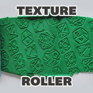 Può includere: Due rulli per texture cilindrici azzurri affiancano una lastra rettangolare verde centrale. I rulli e la lastra verde presentano un motivo in rilievo di varie forme di dadi poliedrici con numeri visibili. È presente il testo TEXTURE ROLLER.