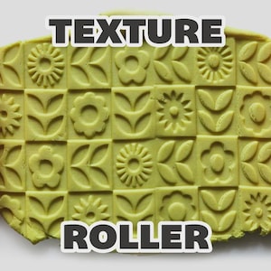 May include: Two orange cylindrical texture rollers with engraved floral and leaf patterns positioned on either side of a light green rectangular slab of clay featuring a repeating grid of embossed flower and leaf designs with the text TEXTURE ROLLER.