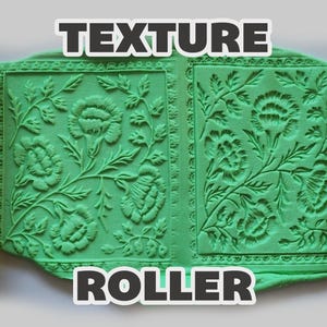 May include: Two yellow cylindrical texture rollers flanking a central green rectangular slab of material. The rollers and the green slab feature an embossed floral pattern with detailed leaves and blossoms. The text TEXTURE ROLLER is visible.