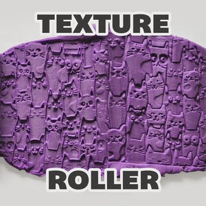 May include: A light green cylindrical texture roller featuring a repeating cat face pattern, shown alongside a purple slab of clay impressed with the same cat design. The text "TEXTURE ROLLER" is displayed in bold white letters above and below the clay.