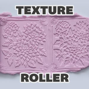 May include: A dark gray cylindrical texture roller with a floral pattern, shown alongside a light purple slab of clay that has been imprinted with the same repeating chrysanthemum flower design. The text TEXTURE ROLLER is visible.